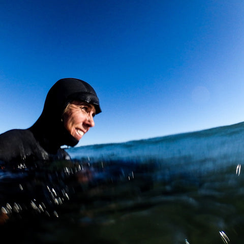 A low-angle close-up of a person's face, wearing a black neoprene hood and smiling slightly, mostly submerged in clear ocean water under a bright, clear blue sky.