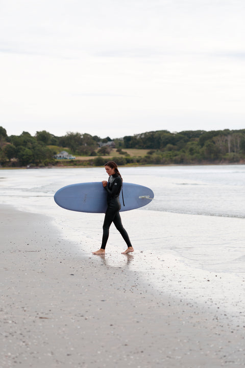 Woman wearing a 00 BY XIIIG PICTA multisport wetsuit walking in from a surf, holding a blue surfboard on a beach with trees in the background