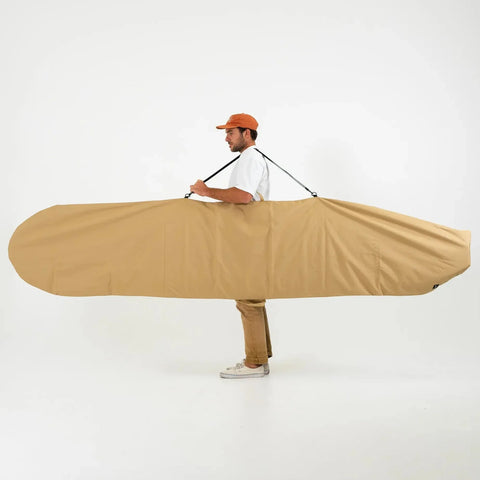 Man carrying a large beige surfboard with a carrying strap on a white background