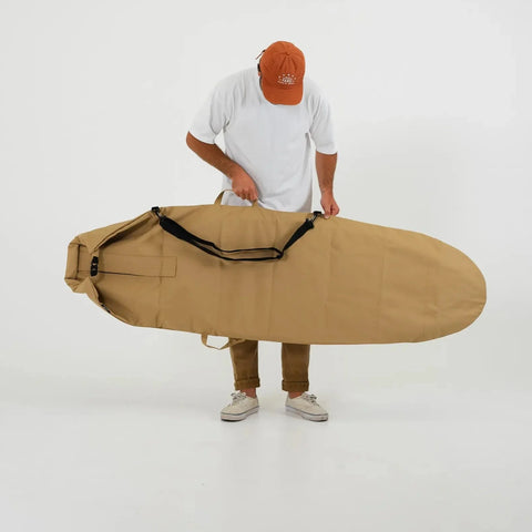 Person holding a tan surfboard bag while putting on the black strap against a white background