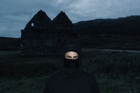 A man wearing a black jacket with a hood pulled over his head and a face mask covering his nose and mouth looks directly at the viewer. Only his eyes are visible. He stands in a dark, desolate landscape with the ruins of a stone building behind him and mountains in the distance under a cloudy sky.