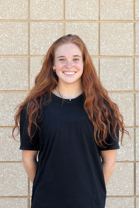 A smiling young woman with long wavy red hair stands in front of a beige tiled wall, wearing a plain black XIIIG short-sleeve cotton t-shirt.