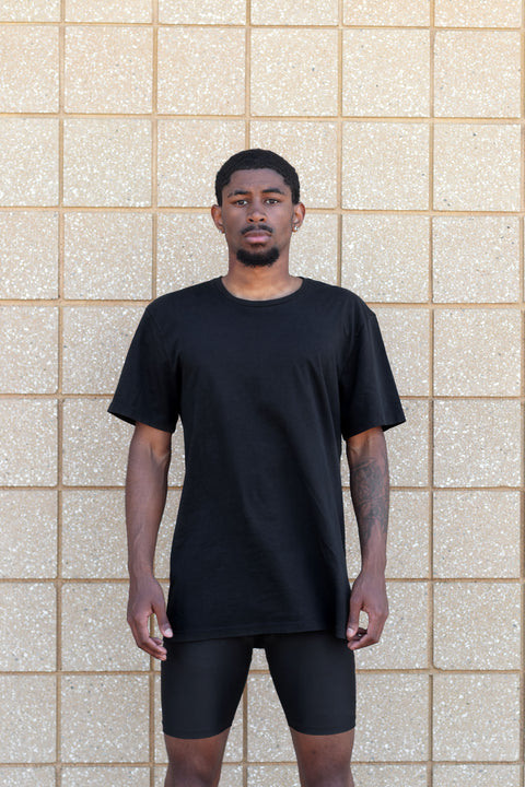 A man wearing a black XIIIG cotton short-sleeve crewneck t-shirt and black XIIIG jammers on, standing in front of a beige tiled wall.